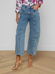 A woman models the Reba Rodeo Jean, a blue high-rise denim wide-leg style featuring front seams, paired with a floral top. She stands in beige heeled sandals against a plain white background. Her head is not visible in the image.