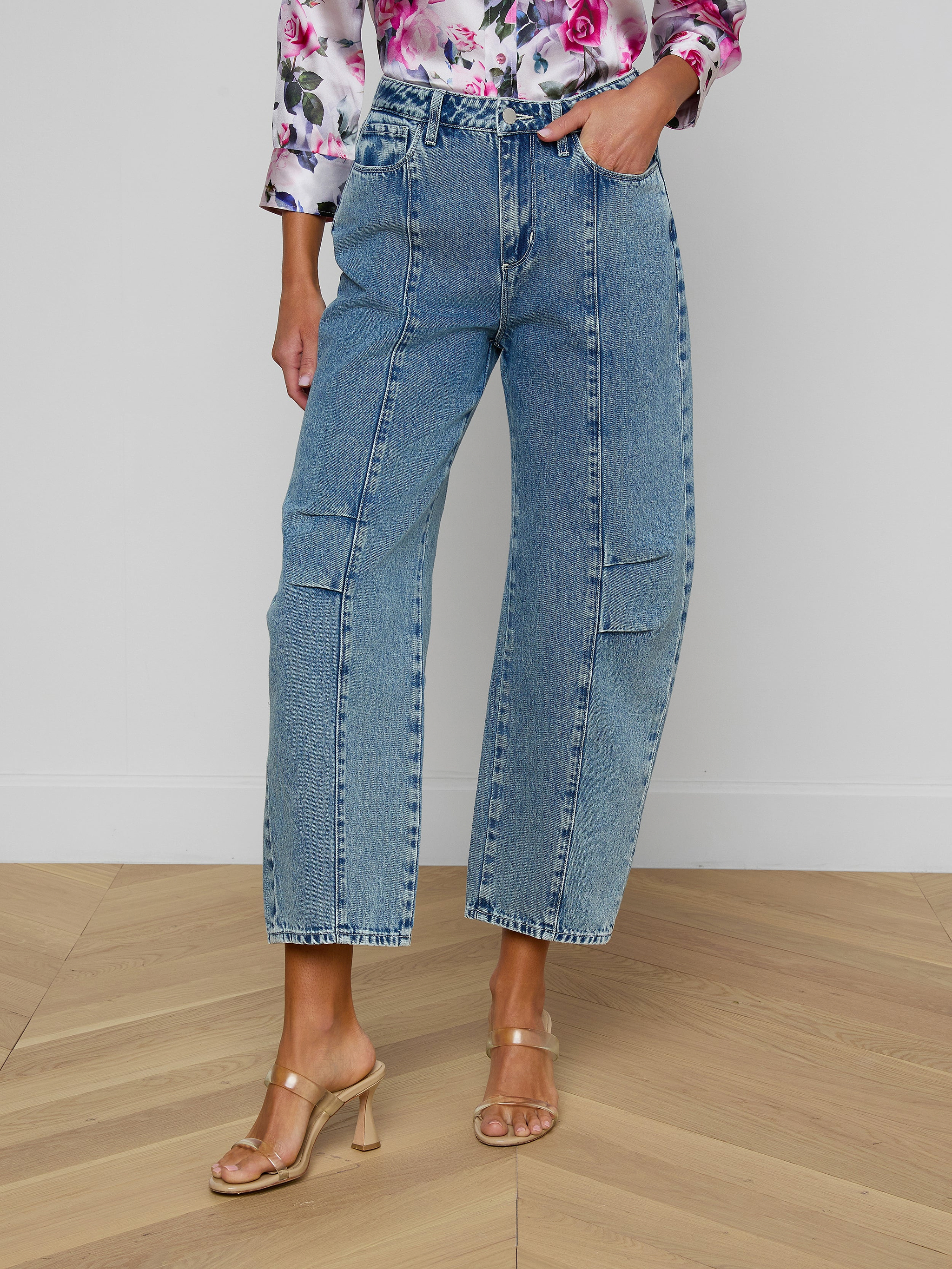 A woman models the Reba Rodeo Jean, a blue high-rise denim wide-leg style featuring front seams, paired with a floral top. She stands in beige heeled sandals against a plain white background. Her head is not visible in the image.