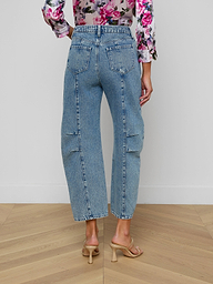 A person is seen from the back wearing the Reba Rodeo Jean in a barrel-leg silhouette with high rise, paired with beige heels. The long-sleeved top features a pink floral pattern, set against a plain light-colored background.