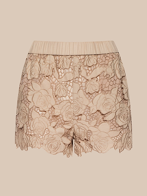 El pantalón corto Laina Eyelet es un pantalón corto de talle alto de color tostado almendrado con un intrincado encaje floral con estampado de rosas y un dobladillo festoneado, que se muestra sobre un fondo beige a juego.