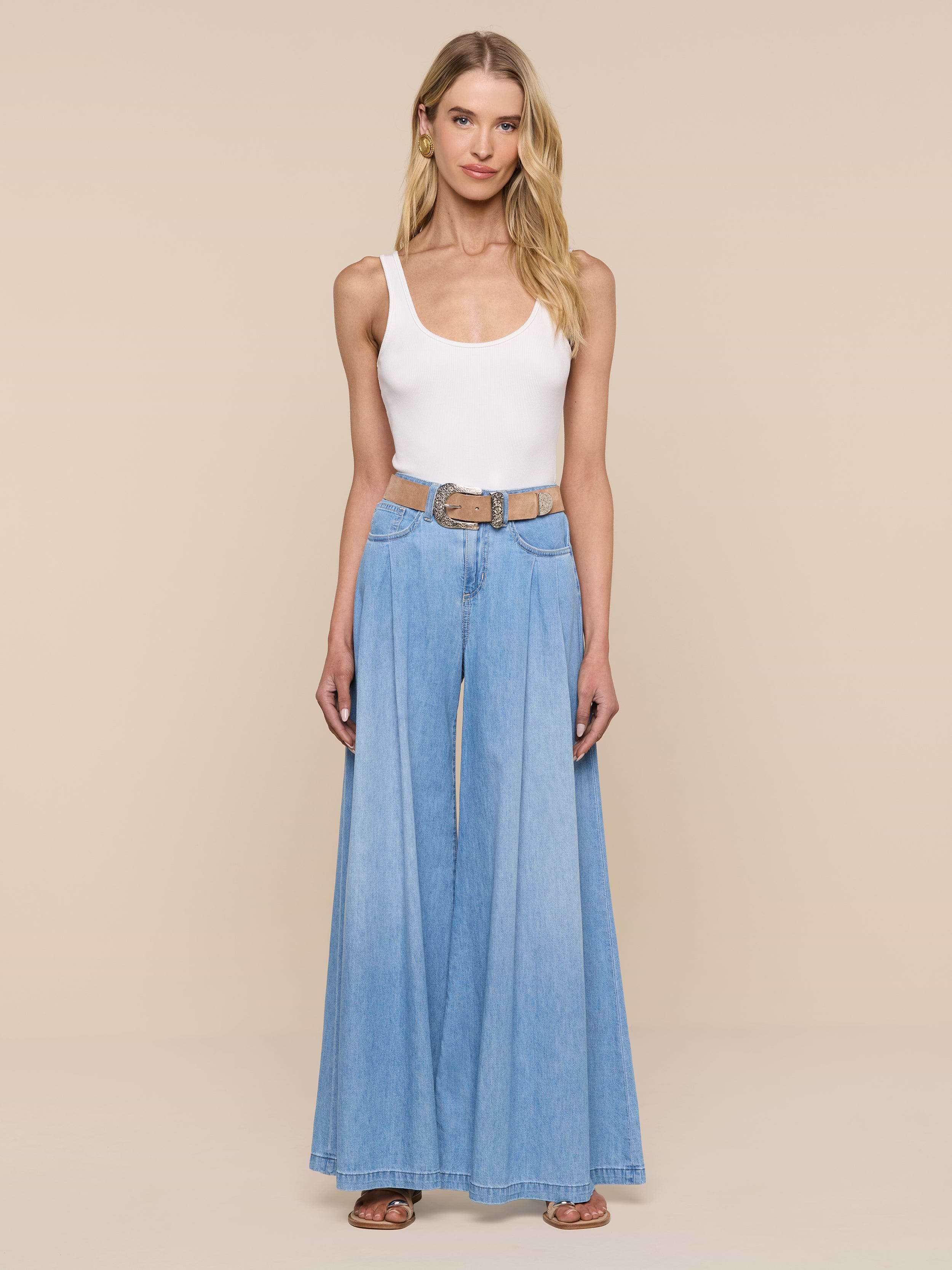 Una donna in piedi, rivolta in avanti su uno sfondo beige, indossa una canottiera bianca e un jeans a gamba larga Dariela con cintura marrone e sandali marroni.