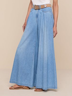 Someone stands against a beige background wearing the Dariela Wide-Leg Jean, featuring a high-waisted, ultra wide leg featherweight denim with front pleats, styled with a tan belt, white top, and tan flat sandals.