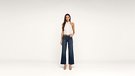 A woman stands against a plain white background, wearing a white halter-neck top, dark wide-leg jeans, a belt, and tan high heels. She has long brown hair and a neutral expression.