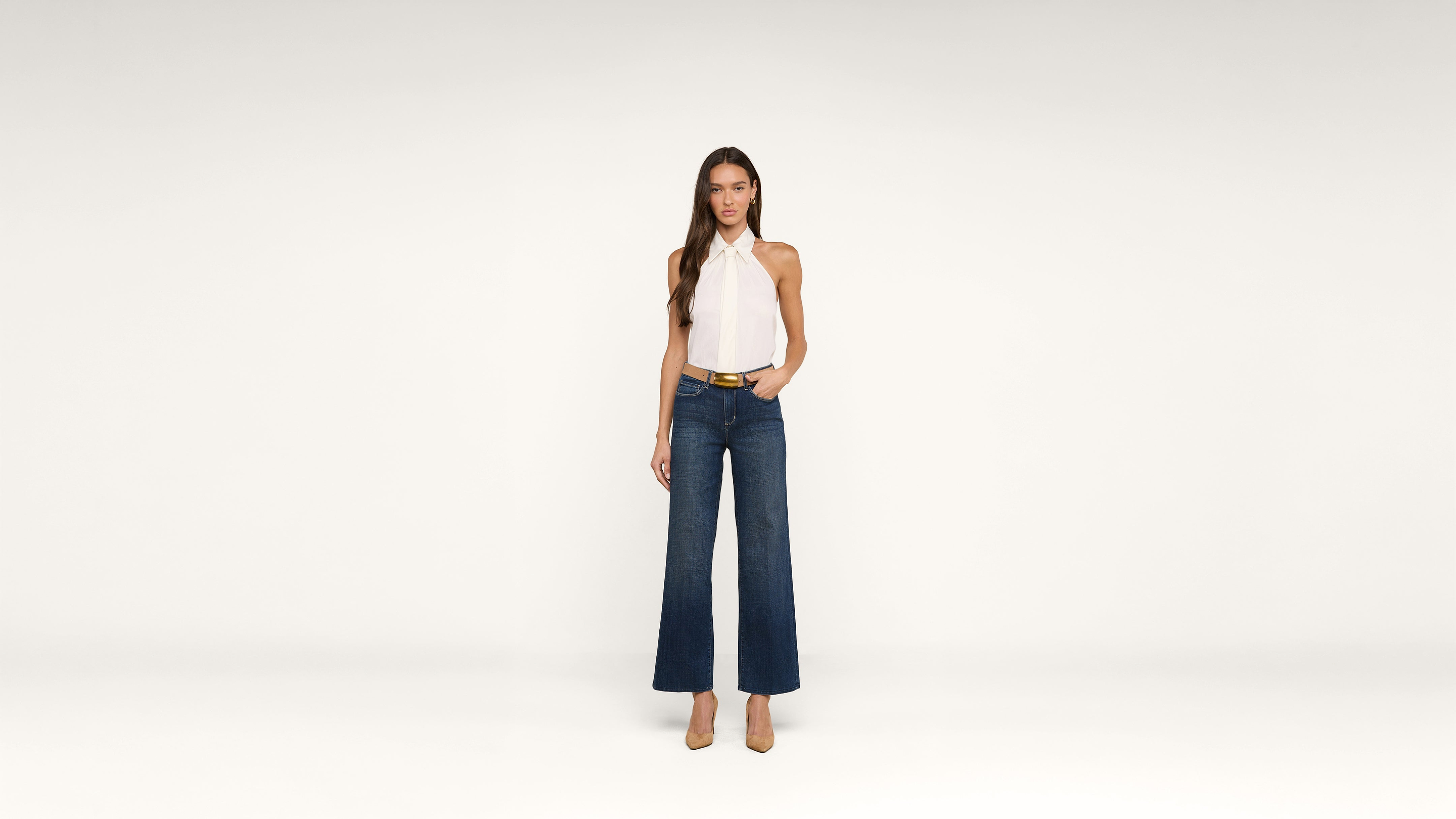 A woman stands against a plain white background, wearing a white halter-neck top, dark wide-leg jeans, a belt, and tan high heels. She has long brown hair and a neutral expression.