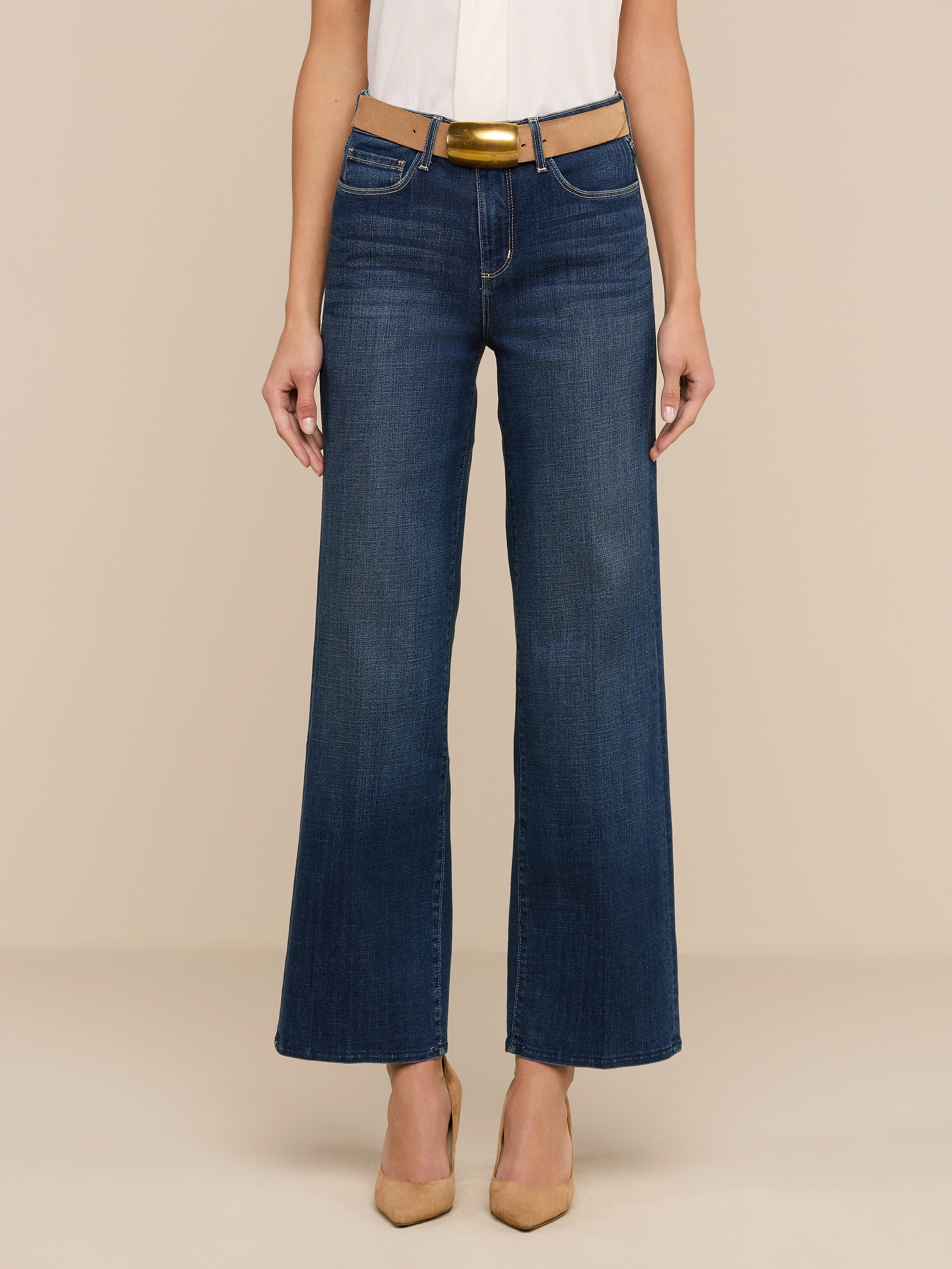 Une personne porte le jean large Rosalyn en denim extensible bleu, associé à une ceinture marron clair, à un haut blanc et à des talons à bouts pointus de couleur beige, sur un fond beige uni.