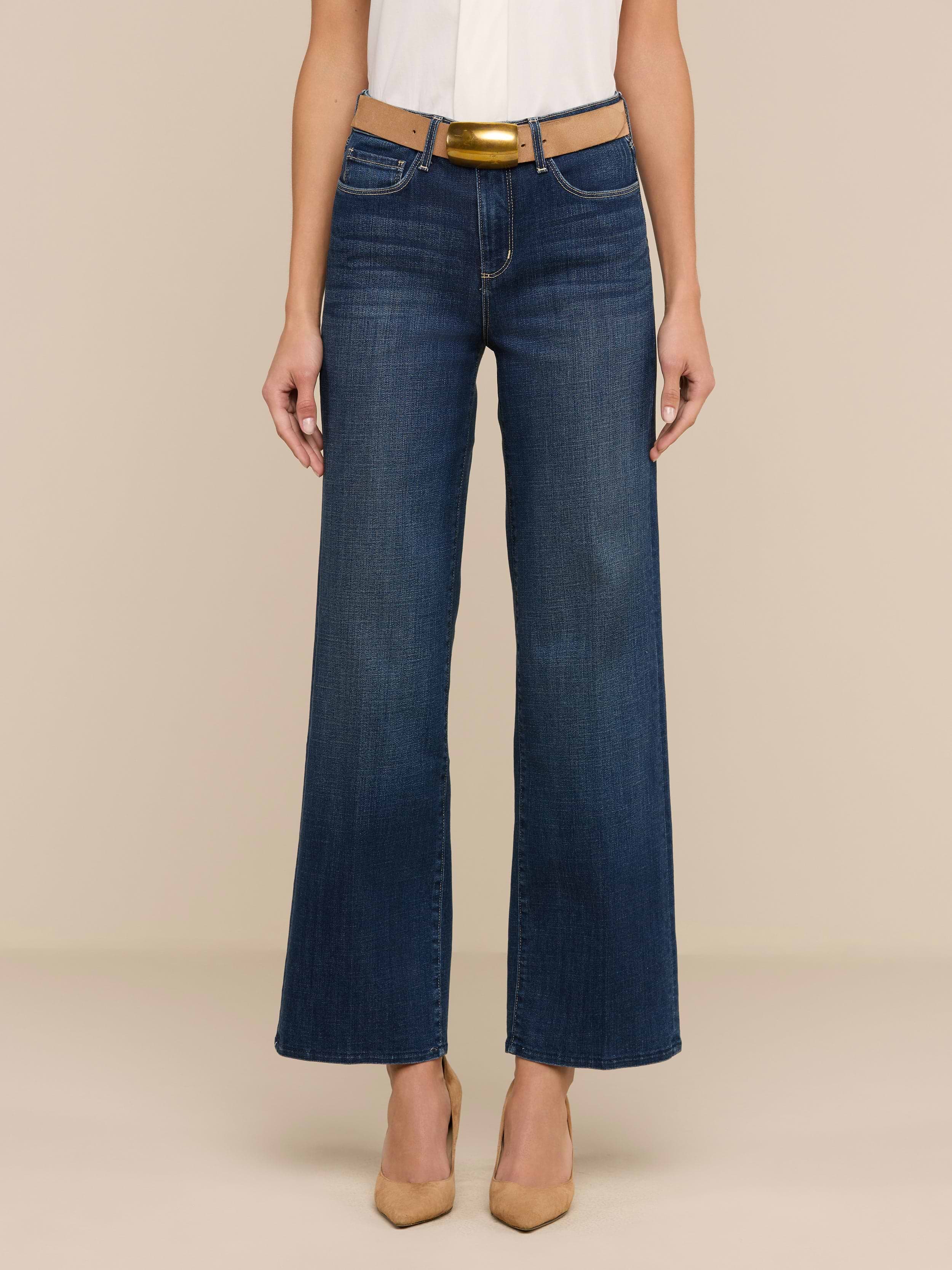 Une personne porte le jean large Rosalyn en denim extensible bleu, associé à une ceinture marron clair, à un haut blanc et à des talons à bouts pointus de couleur beige, sur un fond beige uni.