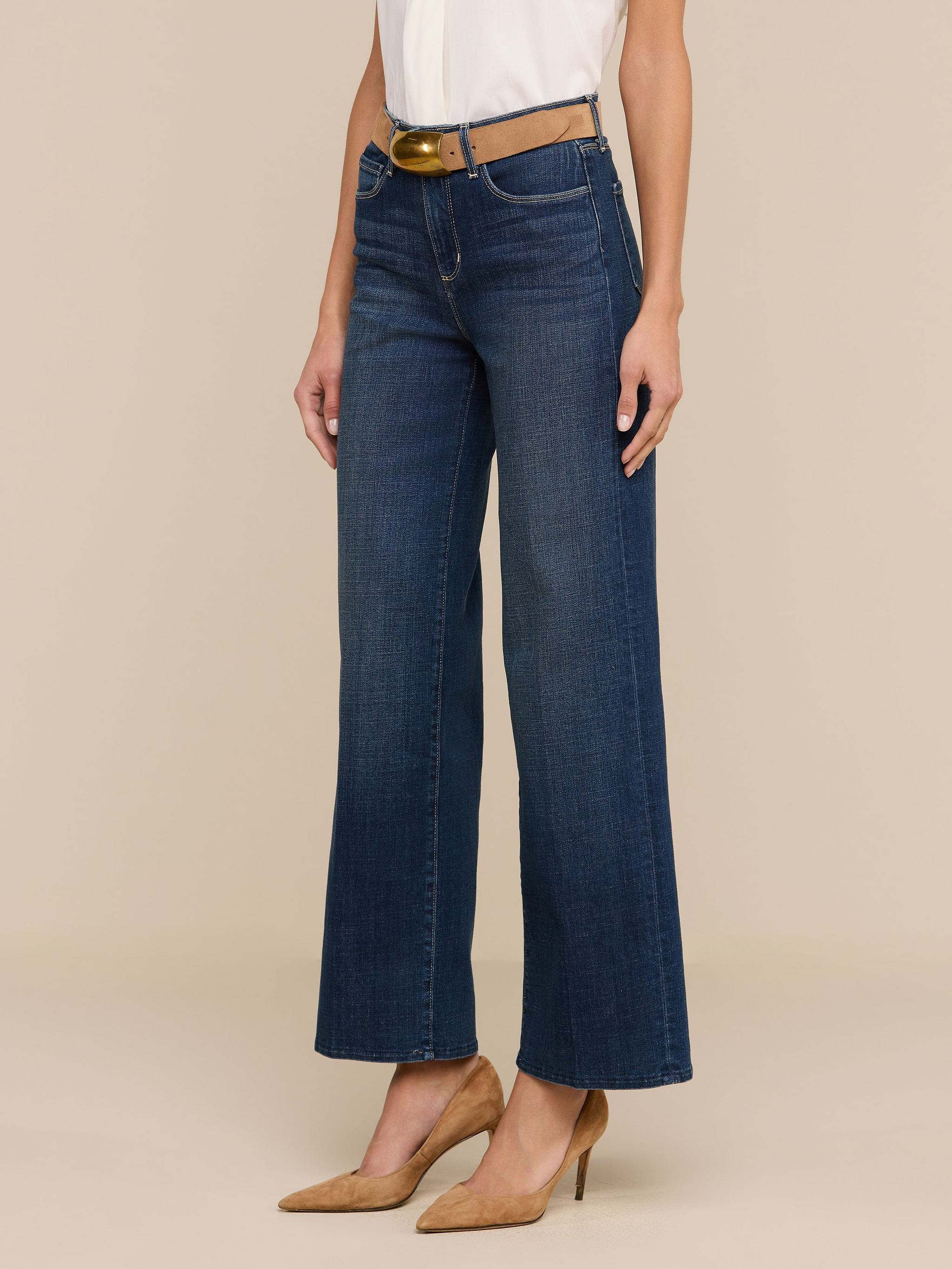 Une personne portant le jean large Rosalyn en denim extensible bleu foncé taille haute, associé à une ceinture marron, un haut blanc rentré dans le pantalon et des chaussures à talons, se tient debout sur un fond beige uni.