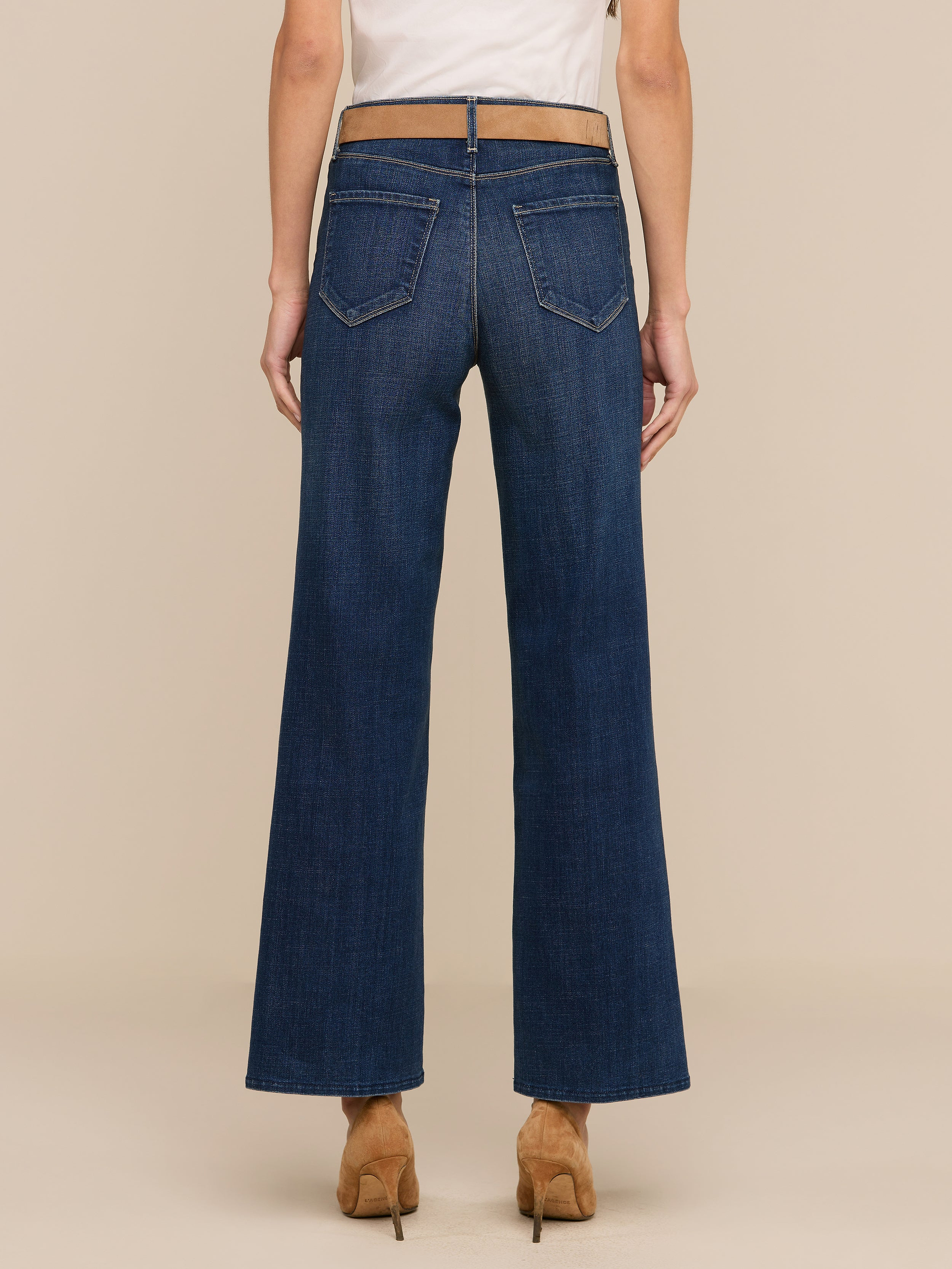 Une personne de face, portant le Rosalyn Wide-Leg Jean en denim extensible bleu foncé à taille haute, avec des poches arrière et des talons de couleur bronze, se tient debout sur un fond beige.