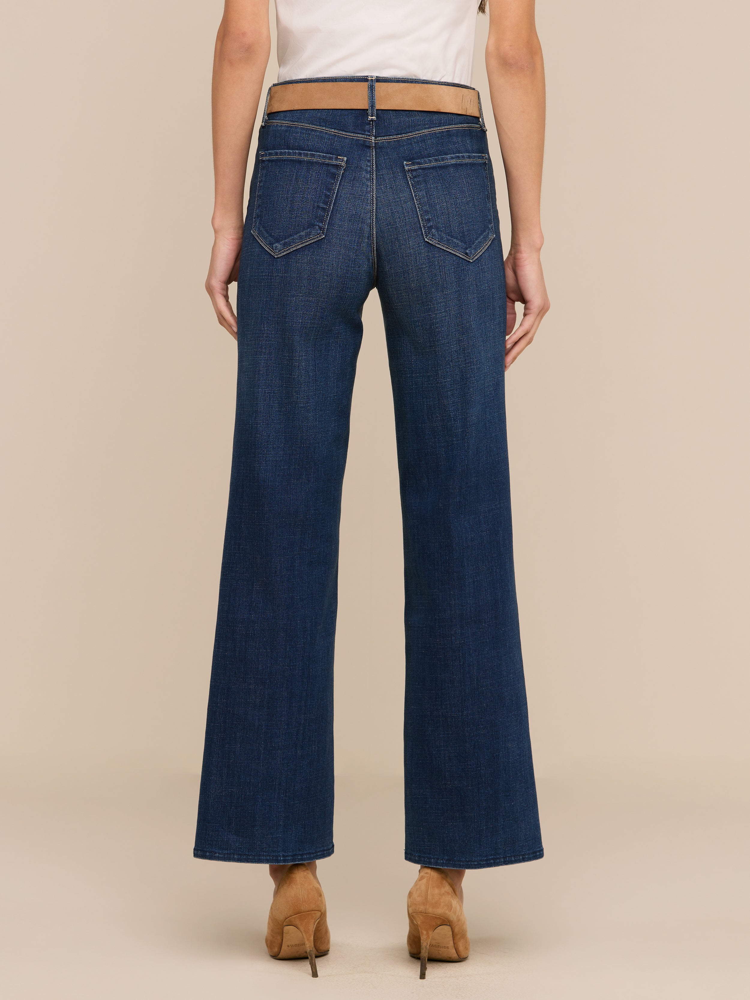 Une personne de face, portant le Rosalyn Wide-Leg Jean en denim extensible bleu foncé à taille haute, avec des poches arrière et des talons de couleur bronze, se tient debout sur un fond beige.