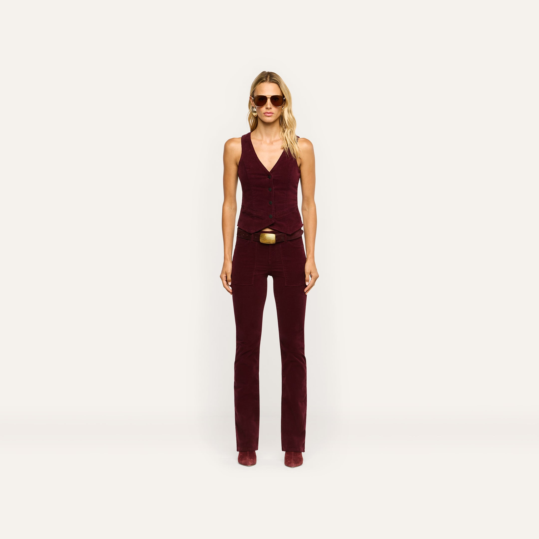 A woman wears a burgundy stretch corduroy vest and matching Saffia Corduroy Pant, a high-rise, slim-straight style featuring a gold-buckled belt.