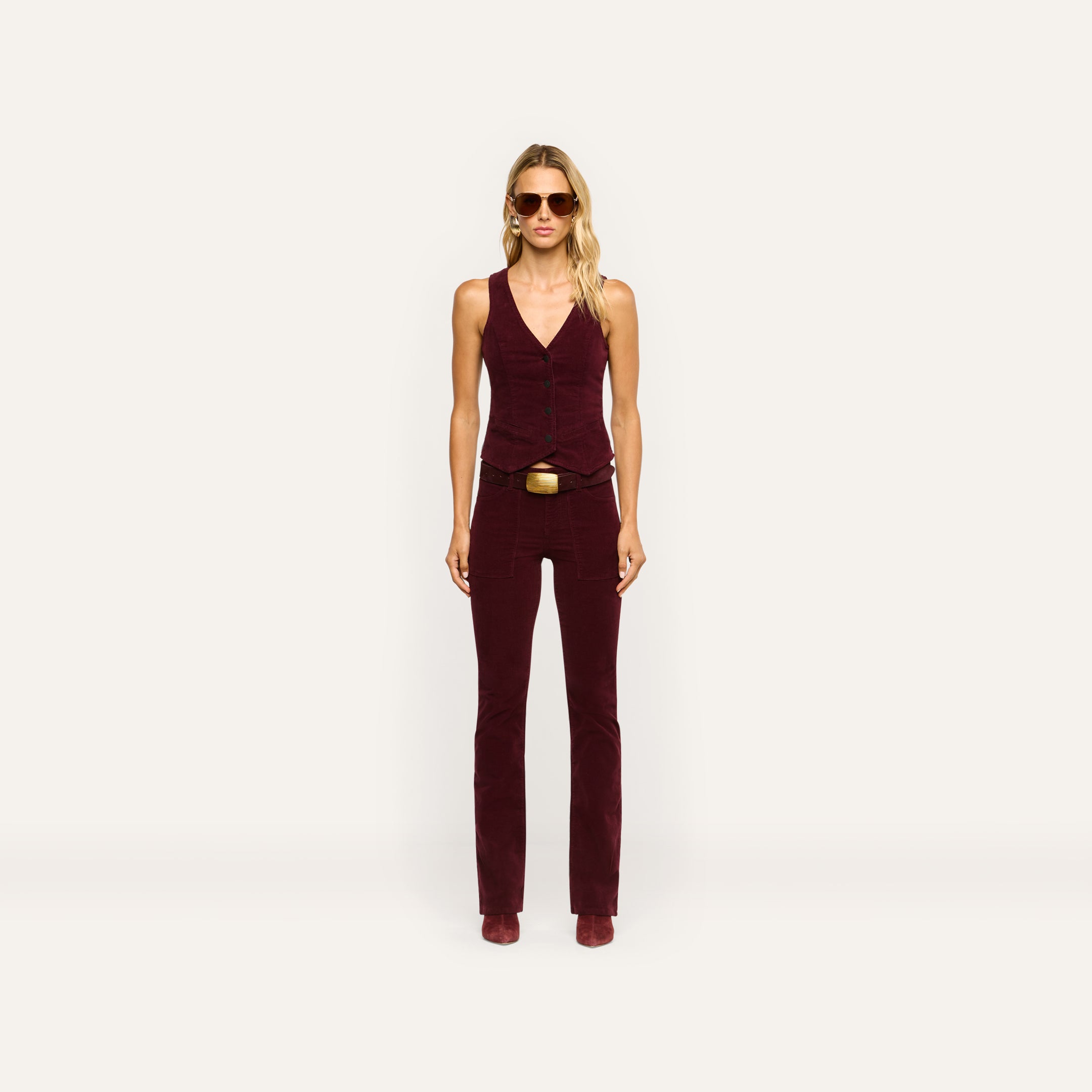 A woman wears a burgundy stretch corduroy vest and matching Saffia Corduroy Pant, a high-rise, slim-straight style featuring a gold-buckled belt.