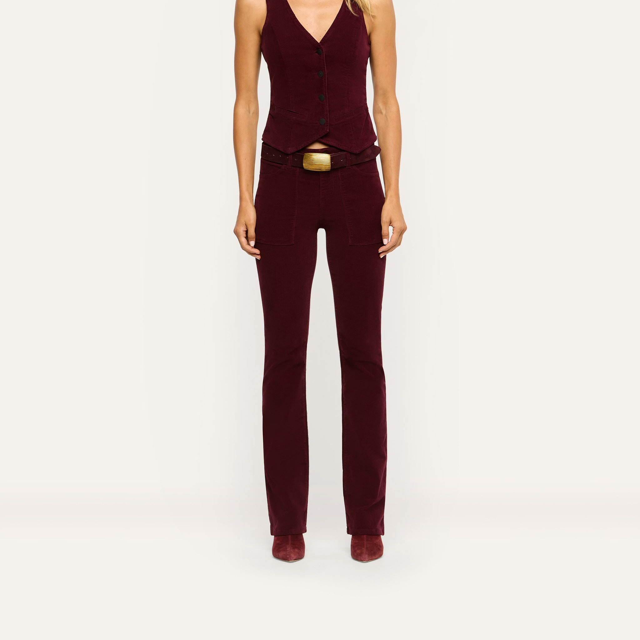 A person wears high-rise burgundy stretch corduroy Saffia Corduroy Pant with a matching button-front vest and a wide brown belt with a large gold buckle.
