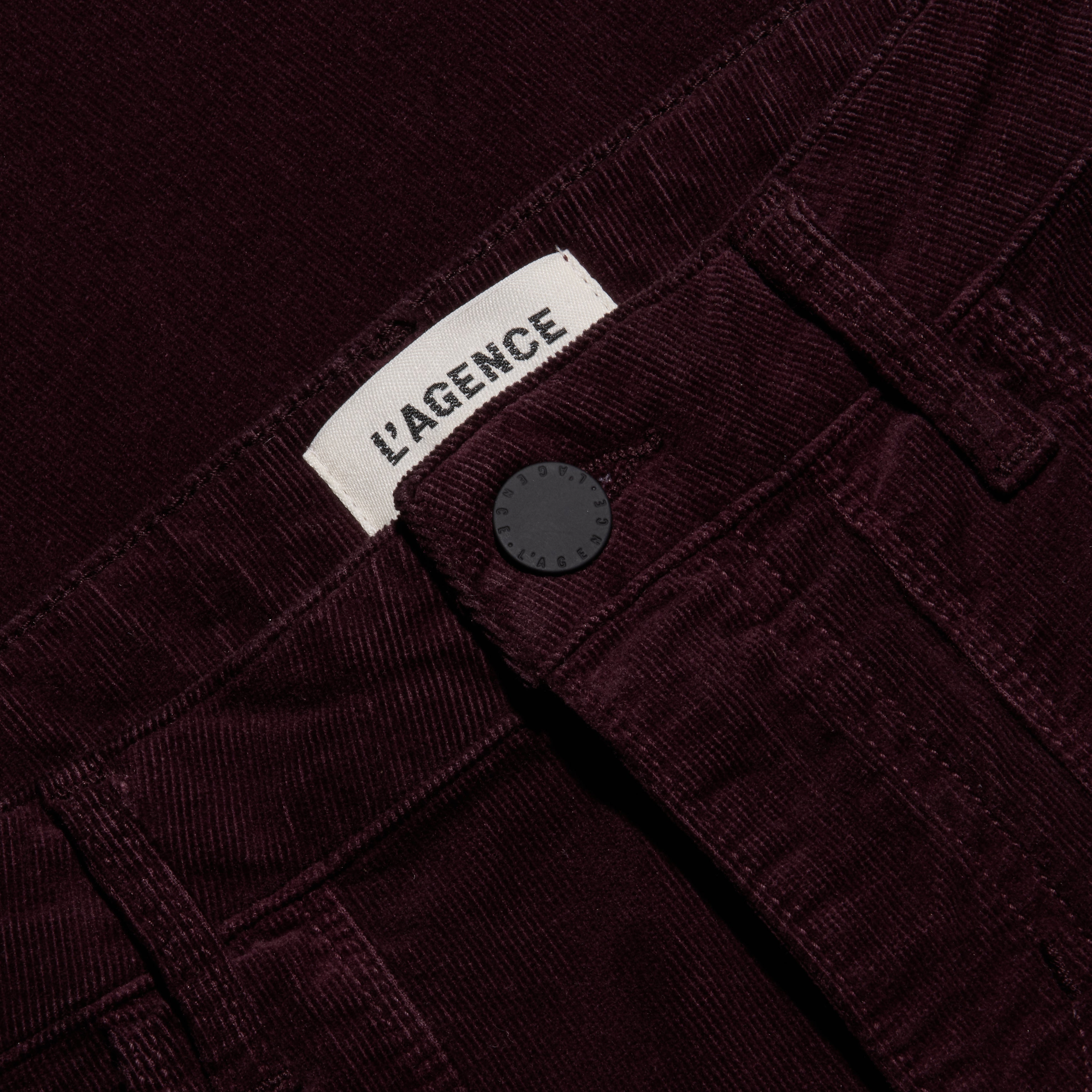Close-up of the waistband of the burgundy Saffia Corduroy Pant, a slim-straight stretch style featuring a button closure and a brand label.