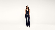 A woman wearing sunglasses, a navy sleeveless top, matching pants, black shoes, and a large belt stands against a plain white background.