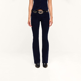 A person stands against a plain white background, wearing a dark blue vest, the Saffia Corduroy Pant with its high-rise, flared silhouette inspired by vintage sophistication, a large decorative belt, and black pointed-toe shoes. Only the body from the shoulders down is visible.