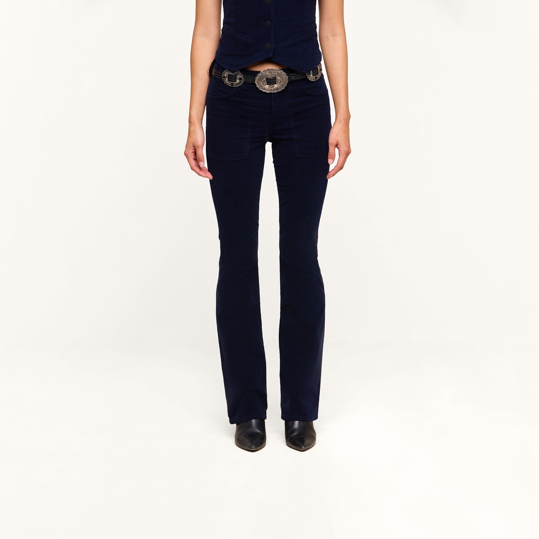 A person wearing the Saffia Corduroy Pant in dark blue, paired with a matching vest, wide belt, and black shoes stands against a plain white background. Their face is not visible.