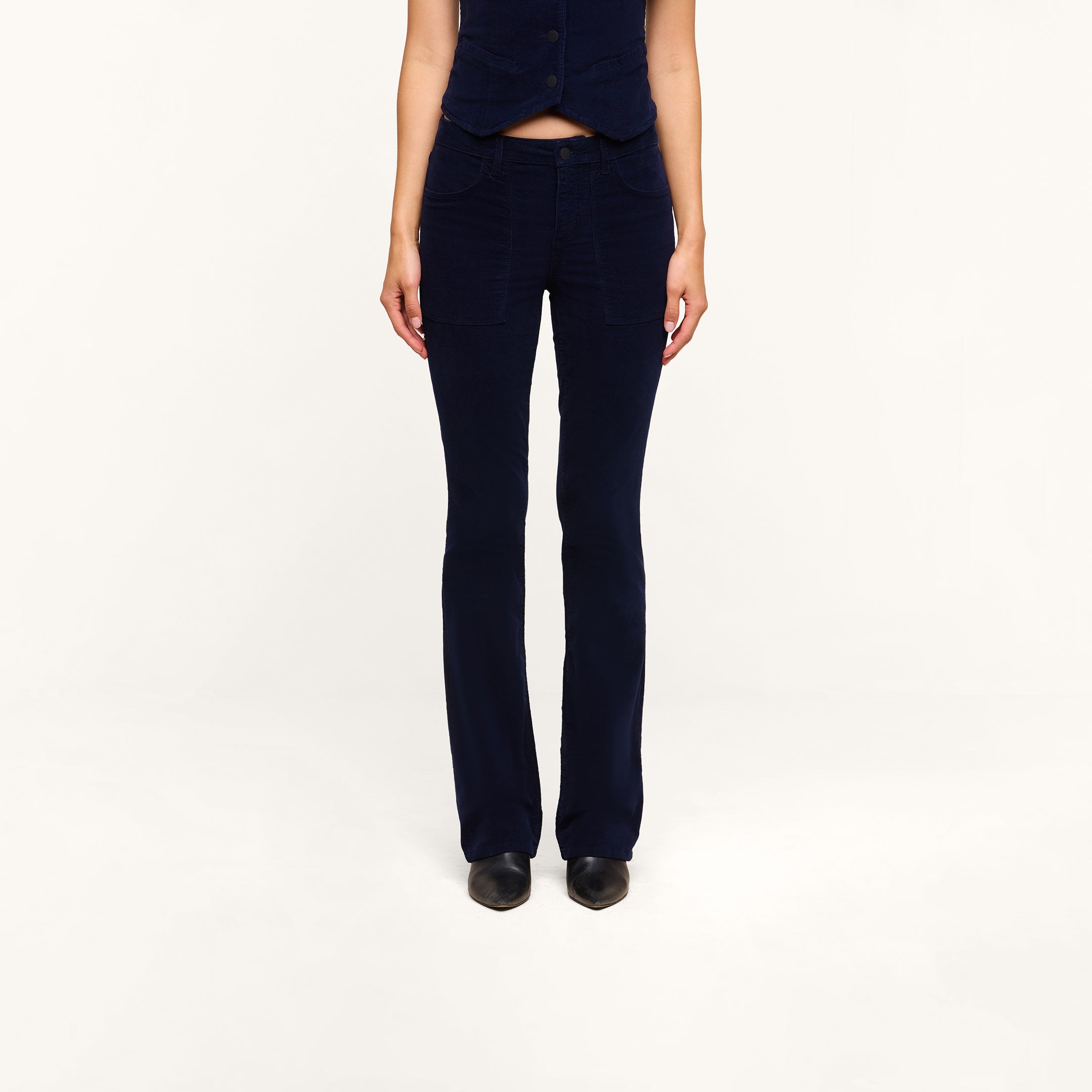 A person stands facing forward with their hands by their sides, wearing the Saffia Corduroy Pant in dark blue—a high-rise, flared style crafted from stretch corduroy—paired with a matching sleeveless vest against a plain white background. Their head is not visible in the image.