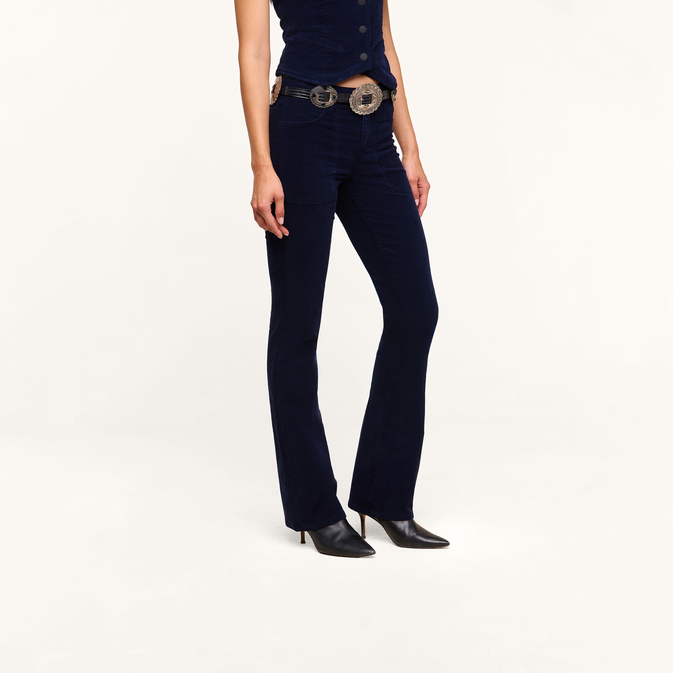 A person wearing fitted dark blue Saffia Corduroy Pants, a matching sleeveless top, a decorative belt with large buckles, and black pointed-toe shoes, standing against a plain white background.