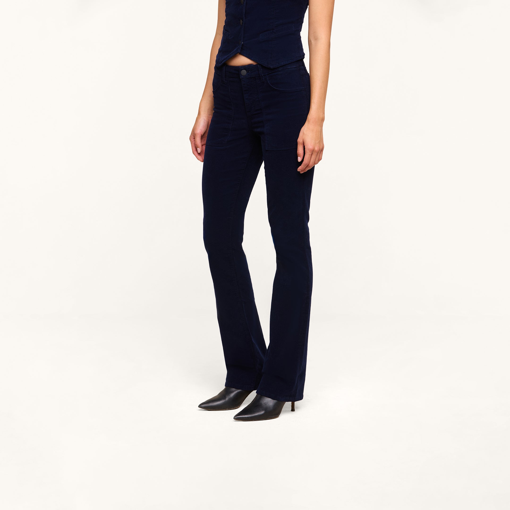 A person wearing the Saffia Corduroy Pant in dark blue—styled as high rise, flared, and crafted from stretch corduroy—paired with a matching sleeveless top and black heeled boots stands against a plain white background. The person’s head is not visible.