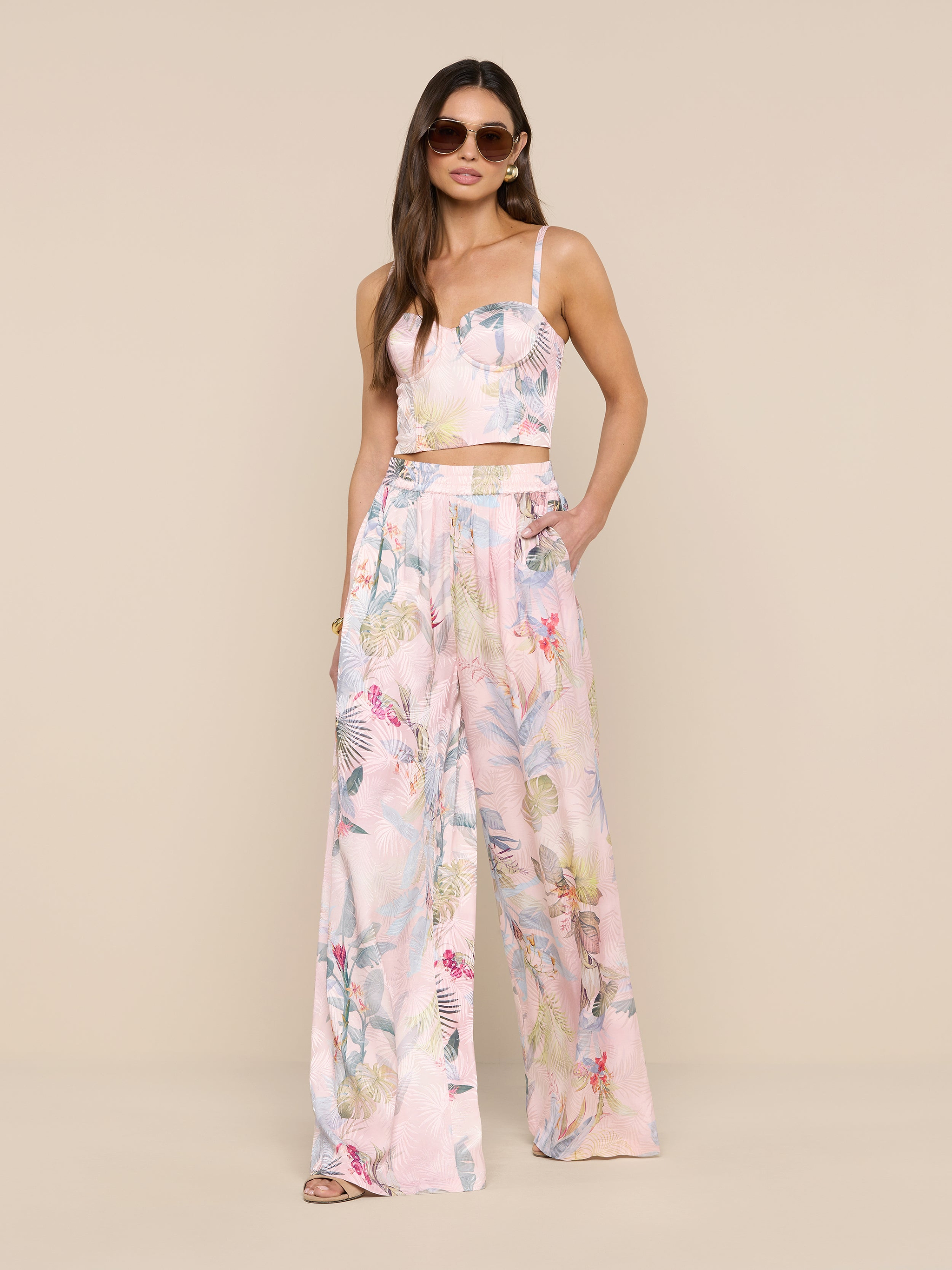 Une femme portant des lunettes de soleil, un top sans manches et un pantalon large en satin Lillian avec un imprimé de feuilles de palmier roses se tient sur un fond beige.