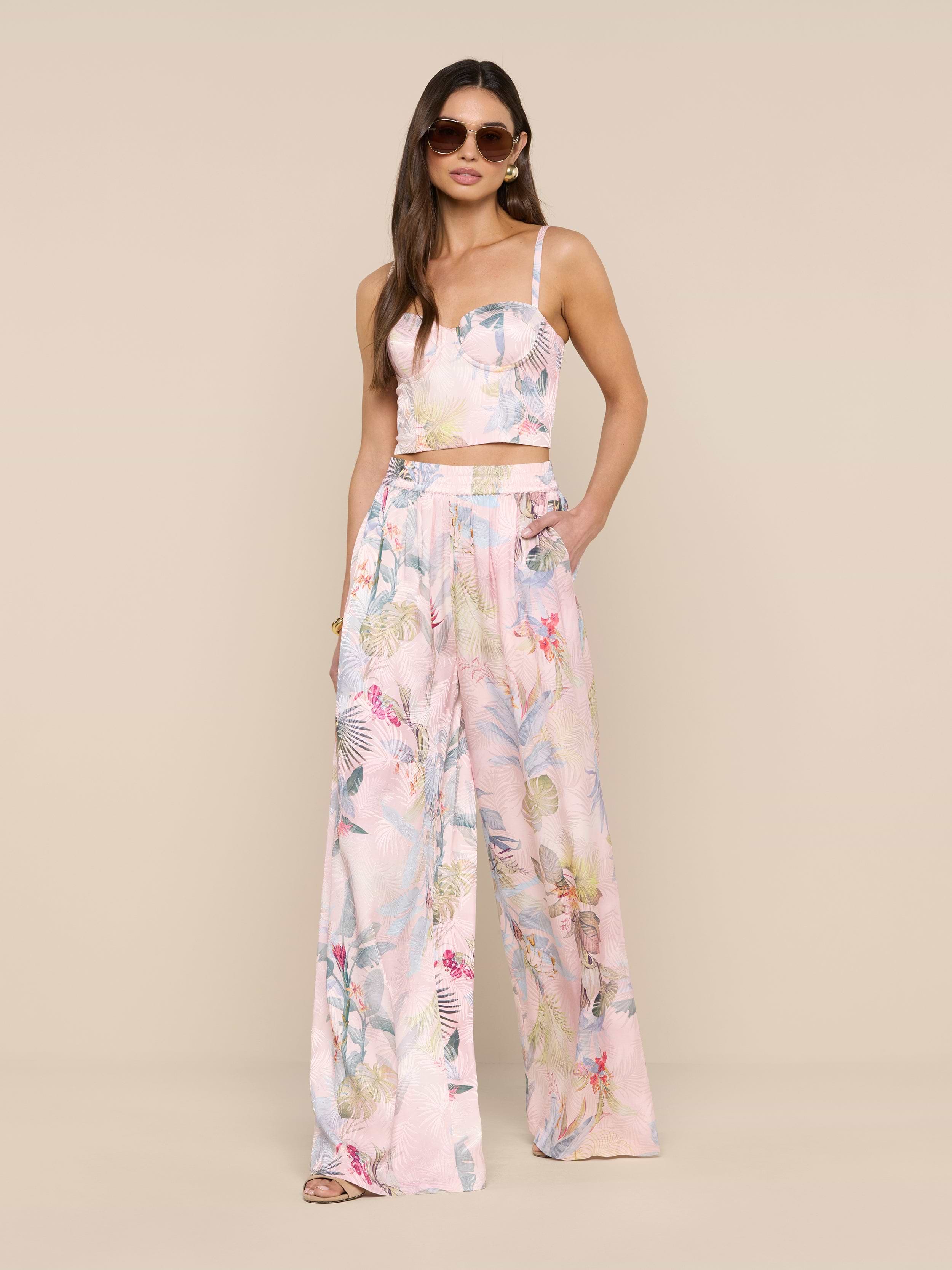 Une femme portant des lunettes de soleil, un top sans manches et un pantalon large en satin Lillian avec un imprimé de feuilles de palmier roses se tient sur un fond beige.
