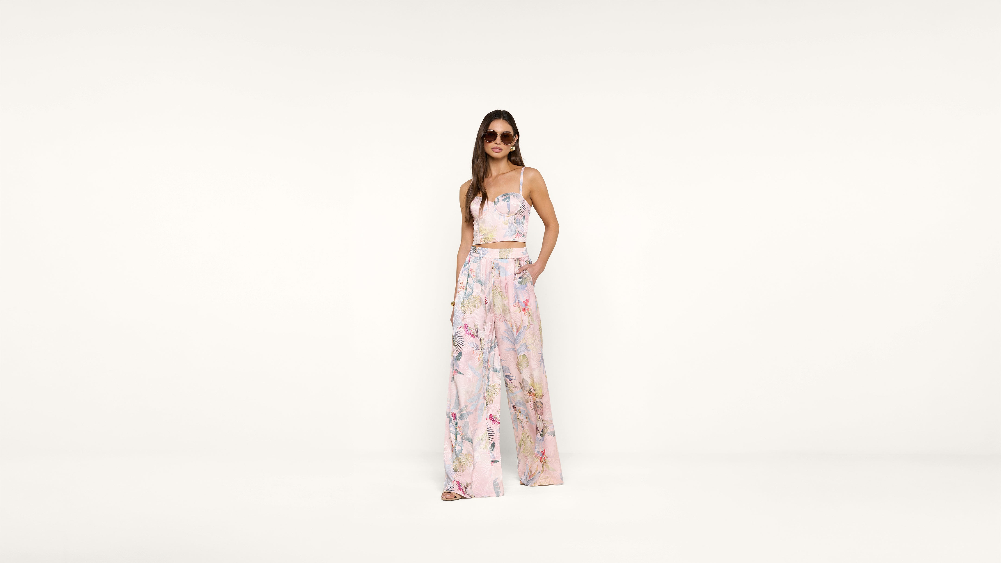 Woman wearing sunglasses and a pastel floral two-piece outfit with a crop top and wide-leg pants, standing against a plain white background.