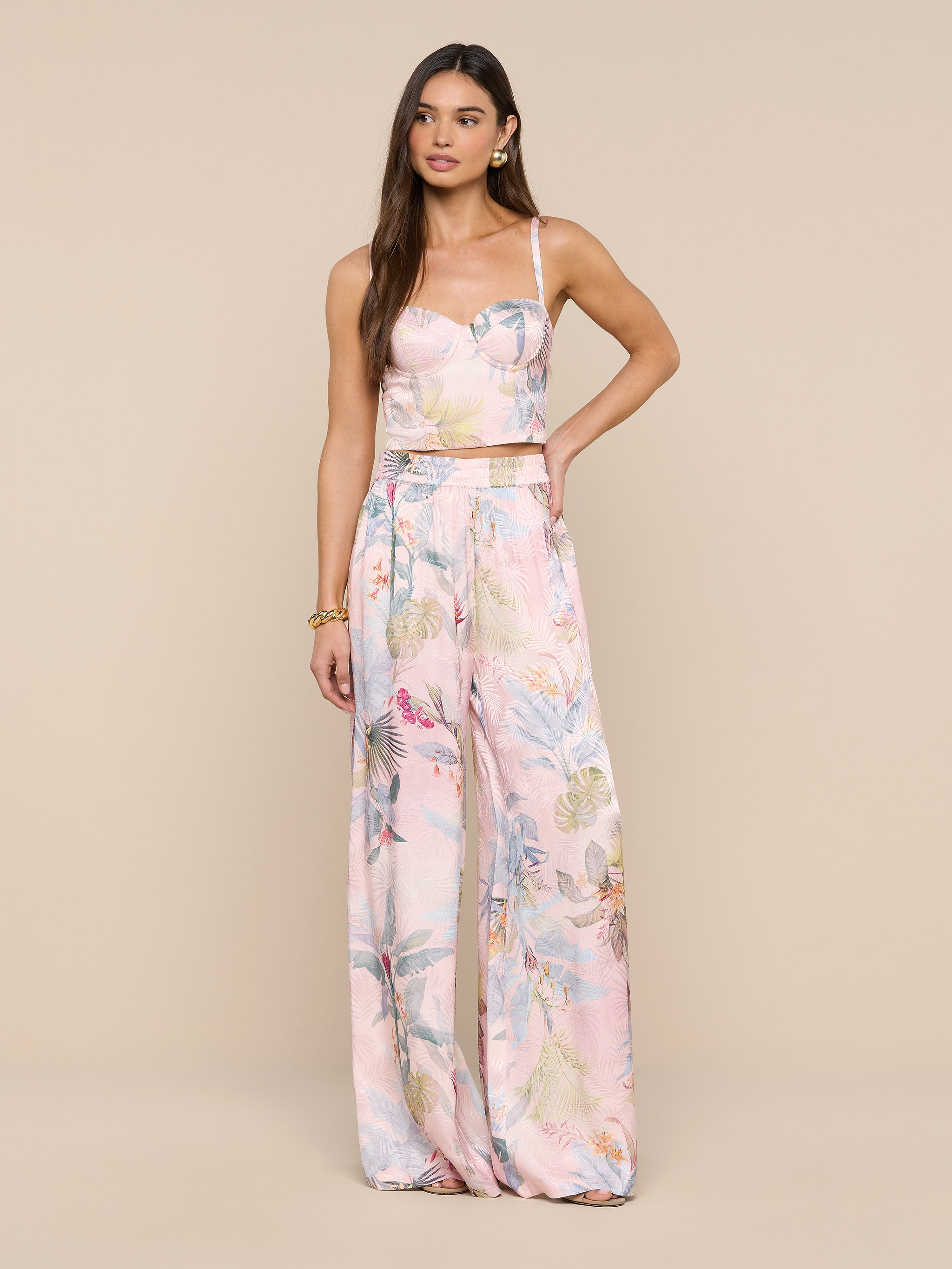 Une femme aux longs cheveux bruns pose sur un fond beige, vêtue dun crop top rose pastel imprimé feuilles de palmier et dun pantalon large en satin Lillian. Elle agrémente son look de bijoux dorés pour une ambiance tropicale chic.