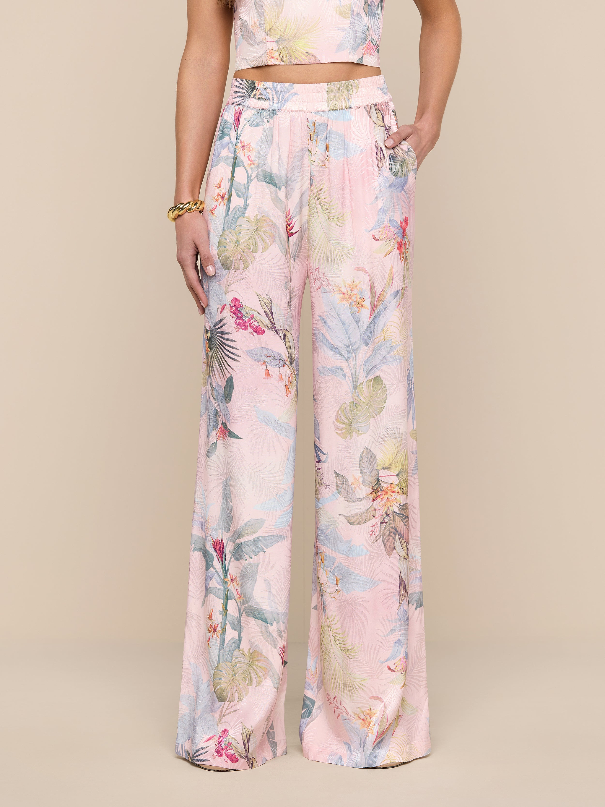 Une personne porte le pantalon large en satin Lillian en jacquard de feuilles de palmier rose, montré de la taille vers le bas sur un fond beige et accessoirisé dun bracelet en or.