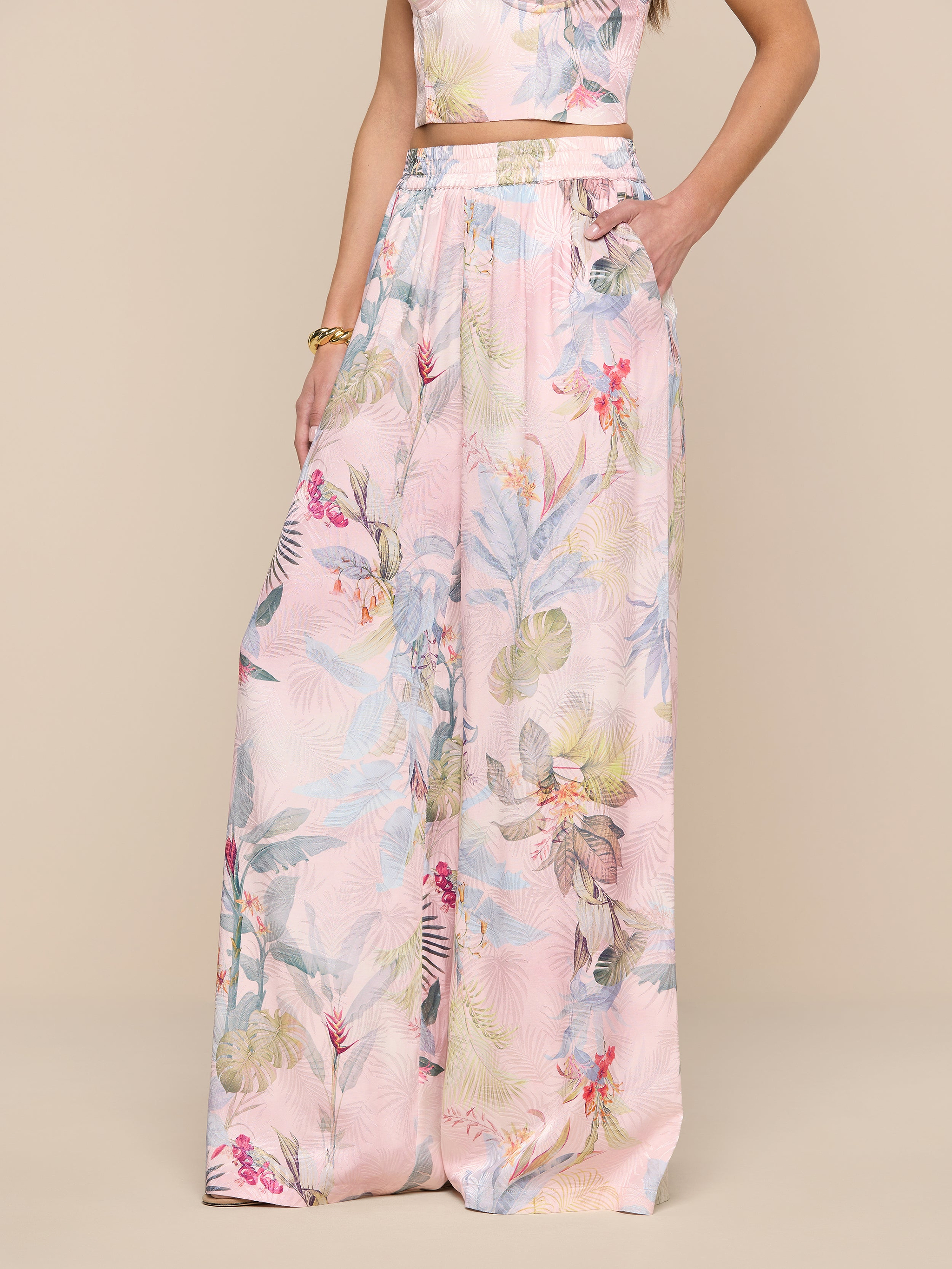 Une femme porte le pantalon à jambes larges Lillian Satin, avec un imprimé de feuilles de palmier roses, une taille haute, une ceinture élastique et des jambes ultra larges ; elle a une main dans sa poche et un crop top assorti est partiellement visible.