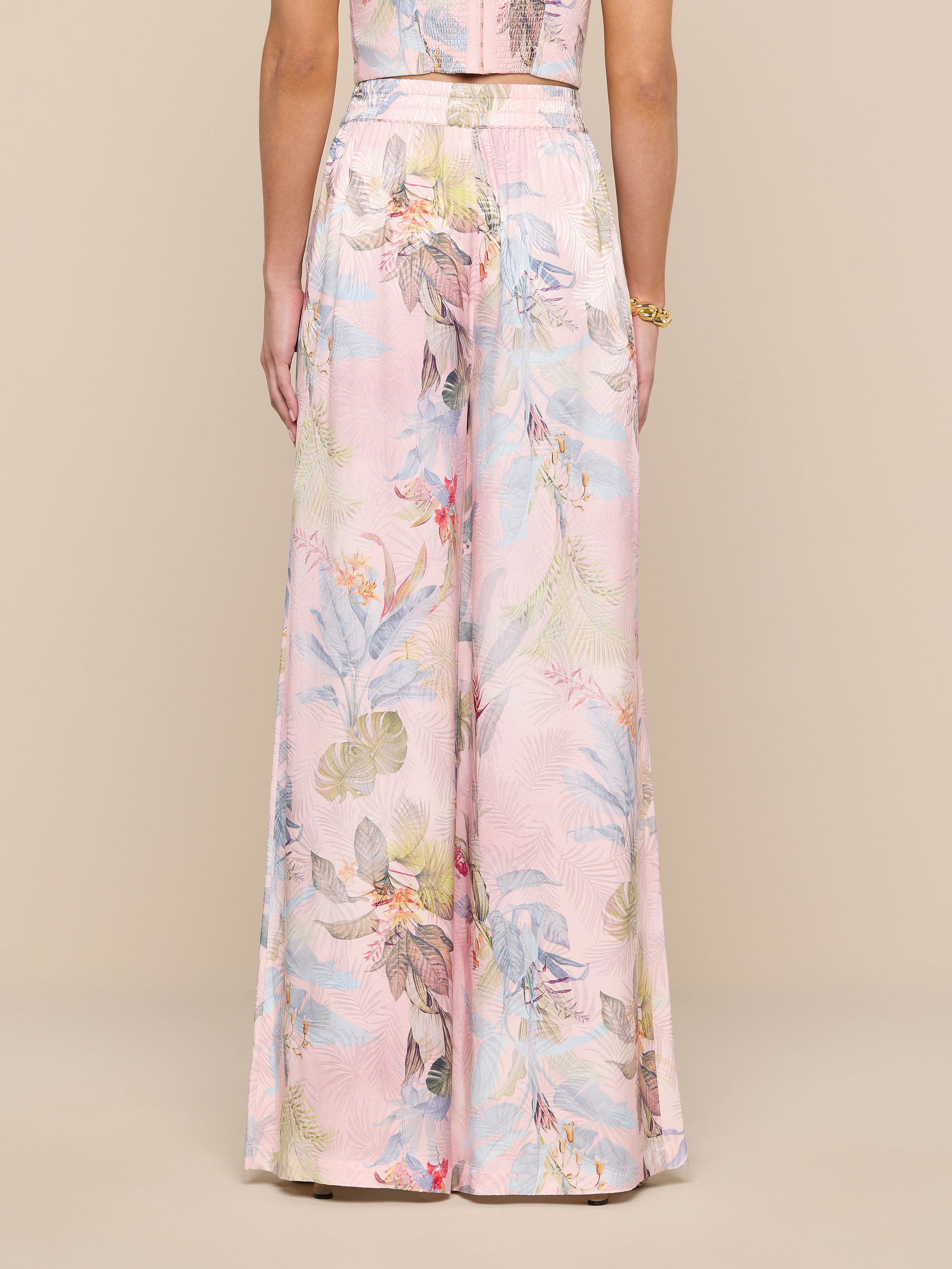Une personne porte le pantalon large en satin Lillian dans un jacquard de feuilles de palmier roses, sur un fond beige. Limage capture une vue de dos de la taille au sol, mettant en valeur la taille haute et la silhouette ultra large.