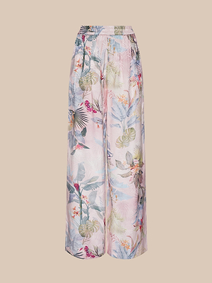 The Lillian Satin Wide-Leg Pant features an ultra-wide silhouette, high elastic waistband, and a pastel pink base adorned with vibrant pink palm leaves and tropical flowers, shown against a plain beige background.