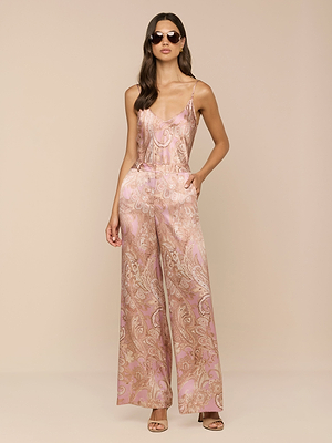 A woman with long, wavy brown hair stands against a beige background wearing sunglasses, nude heeled sandals, and the Pilar Silk Wide-Leg Pant in a pink and gold paisley print. She has her hands in her pockets.