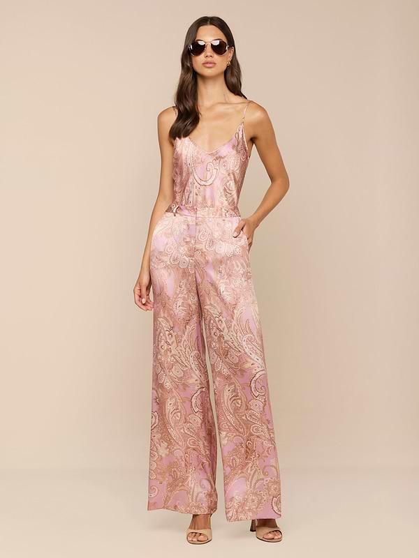 Une femme présente le pantalon Pilar en soie à jambe large, avec un imprimé cachemire rose et une coupe à taille haute. Elle associe cet élégant pantalon en satin à des lunettes de soleil et à des talons ouverts, posant avec assurance sur un fond clair et uni.