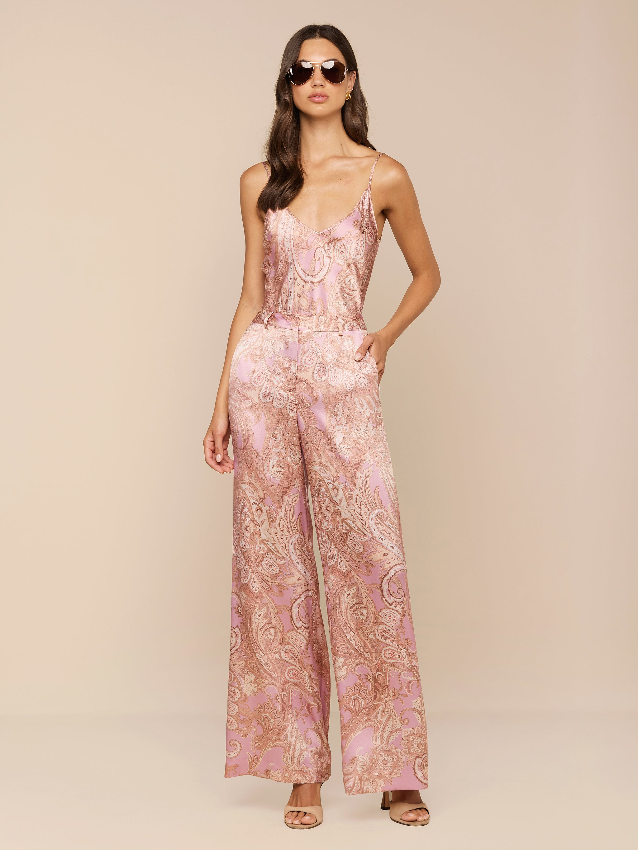 Une femme présente le pantalon Pilar en soie à jambe large, avec un imprimé cachemire rose et une coupe à taille haute. Elle associe cet élégant pantalon en satin à des lunettes de soleil et à des talons ouverts, posant avec assurance sur un fond clair et uni.