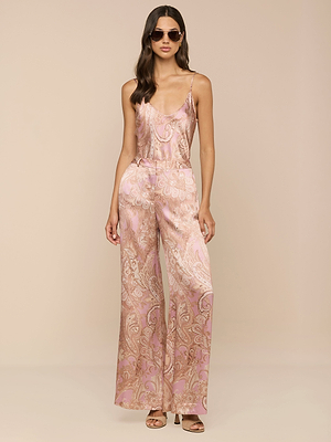 A woman wearing sunglasses and heeled sandals stands against a beige background in silky, high-rise Pilar Silk Wide-Leg Pants with a pink paisley print, her hands in her pockets.