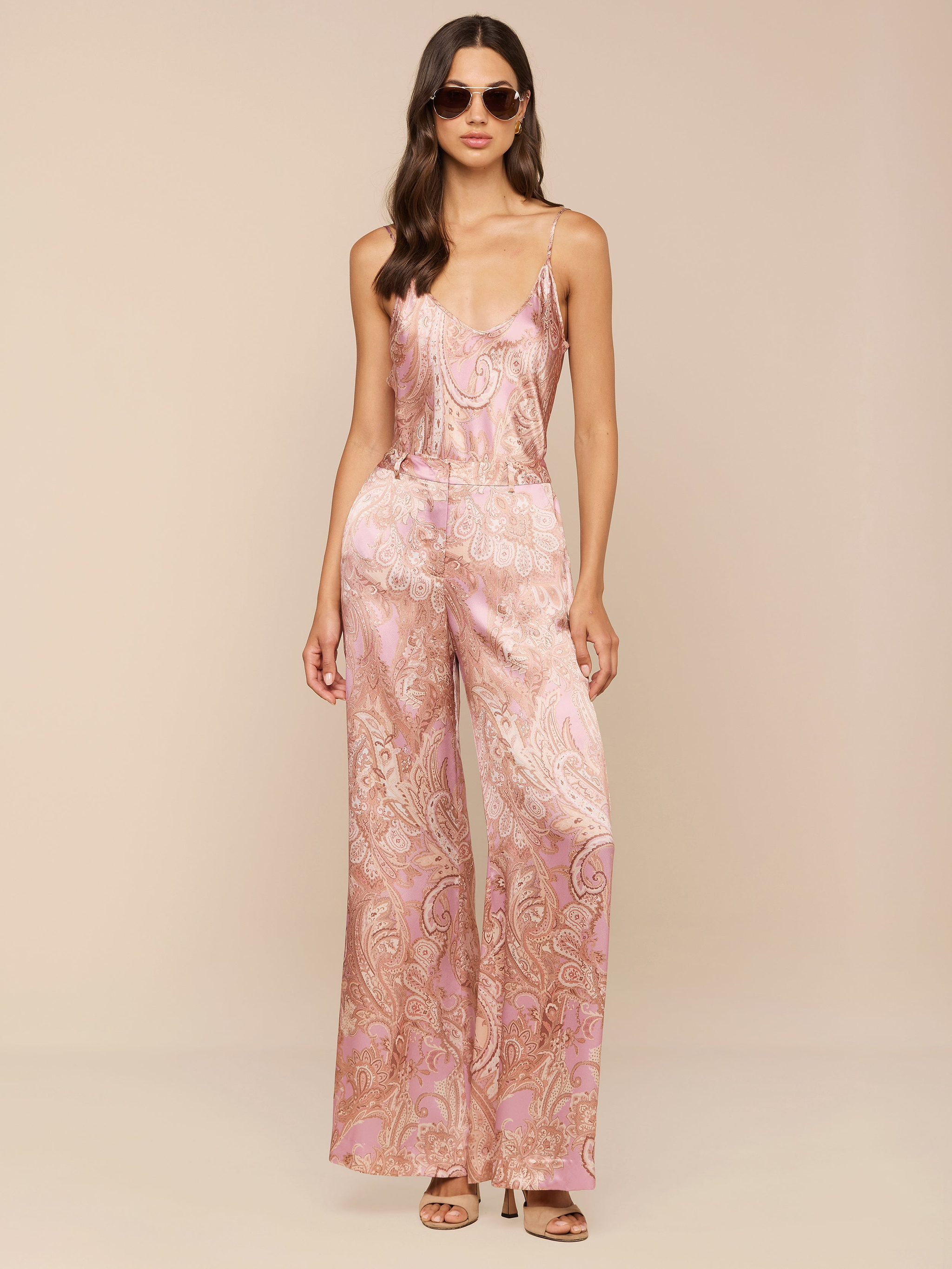 Une femme pose avec assurance sur un fond neutre, portant des lunettes de soleil et le pantalon large en soie Pilar à imprimé paisley mauve, avec des bretelles spaghetti pour un look chic.