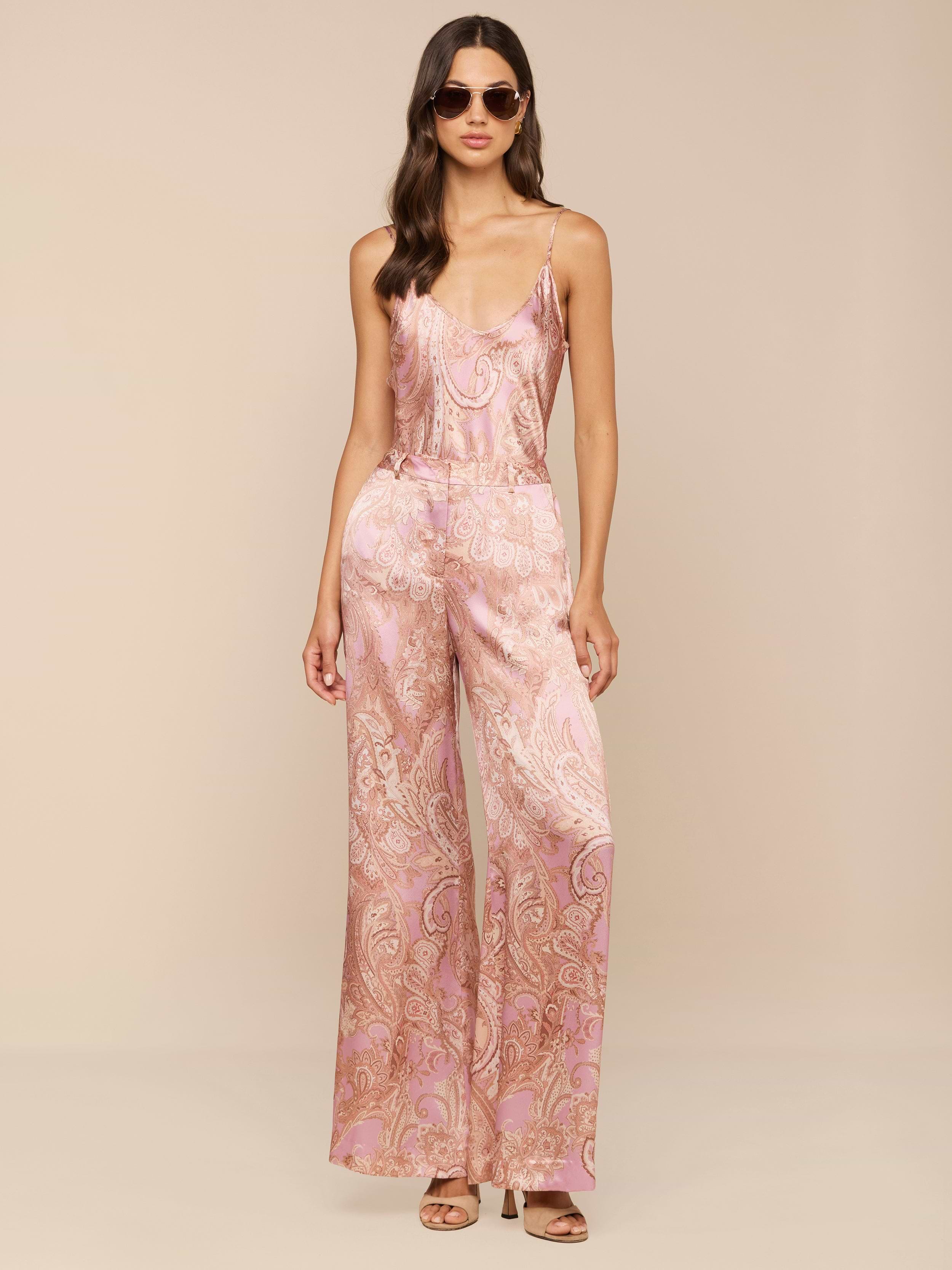 Une femme pose avec assurance sur un fond neutre, portant des lunettes de soleil et le pantalon large en soie Pilar à imprimé paisley mauve, avec des bretelles spaghetti pour un look chic.