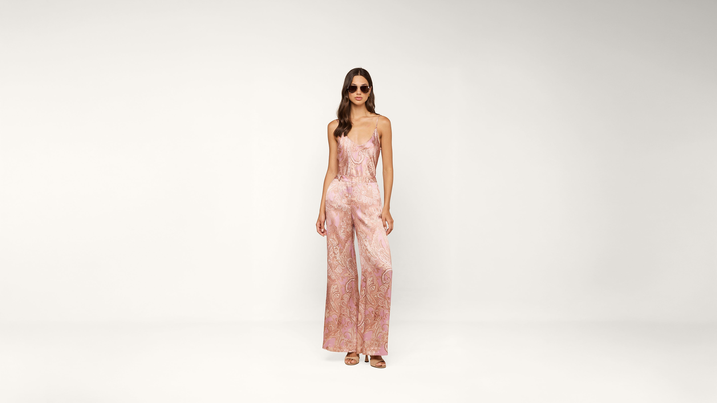 A woman wearing sunglasses and a pink patterned sleeveless jumpsuit stands against a plain white background.
