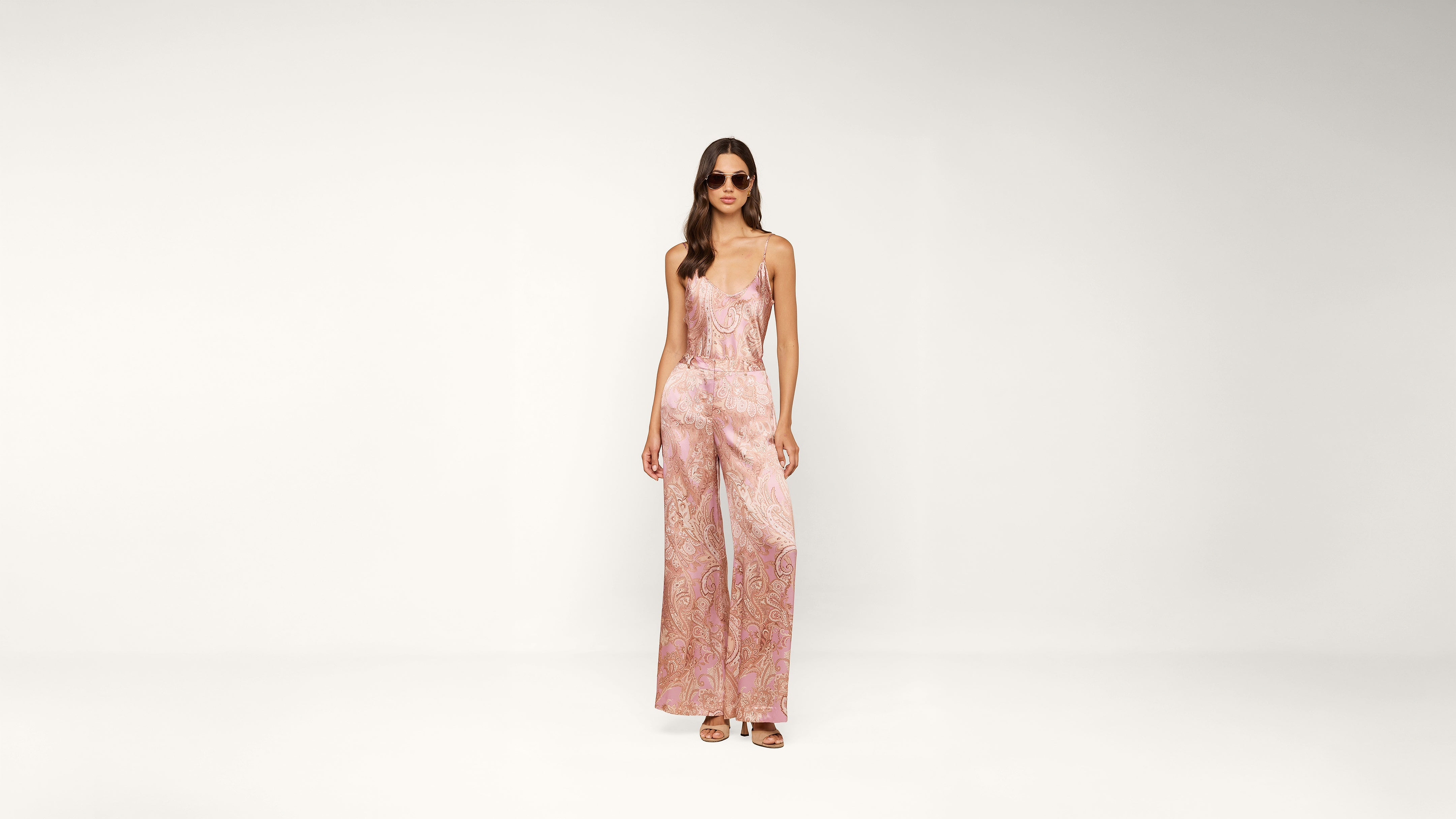 A woman wearing sunglasses and a pink patterned sleeveless jumpsuit stands against a plain white background.