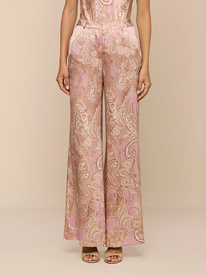 A woman stands against a neutral background wearing Pilar Silk Wide-Leg Pants featuring a pink and beige paisley print, paired with open-toe heels.