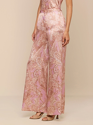 Portant le pantalon large Pilar en soie à imprimé cachemire rose et beige, associé à des talons nude ouverts, une personne se tient debout sur un fond beige, le haut de son corps étant partiellement hors champ.