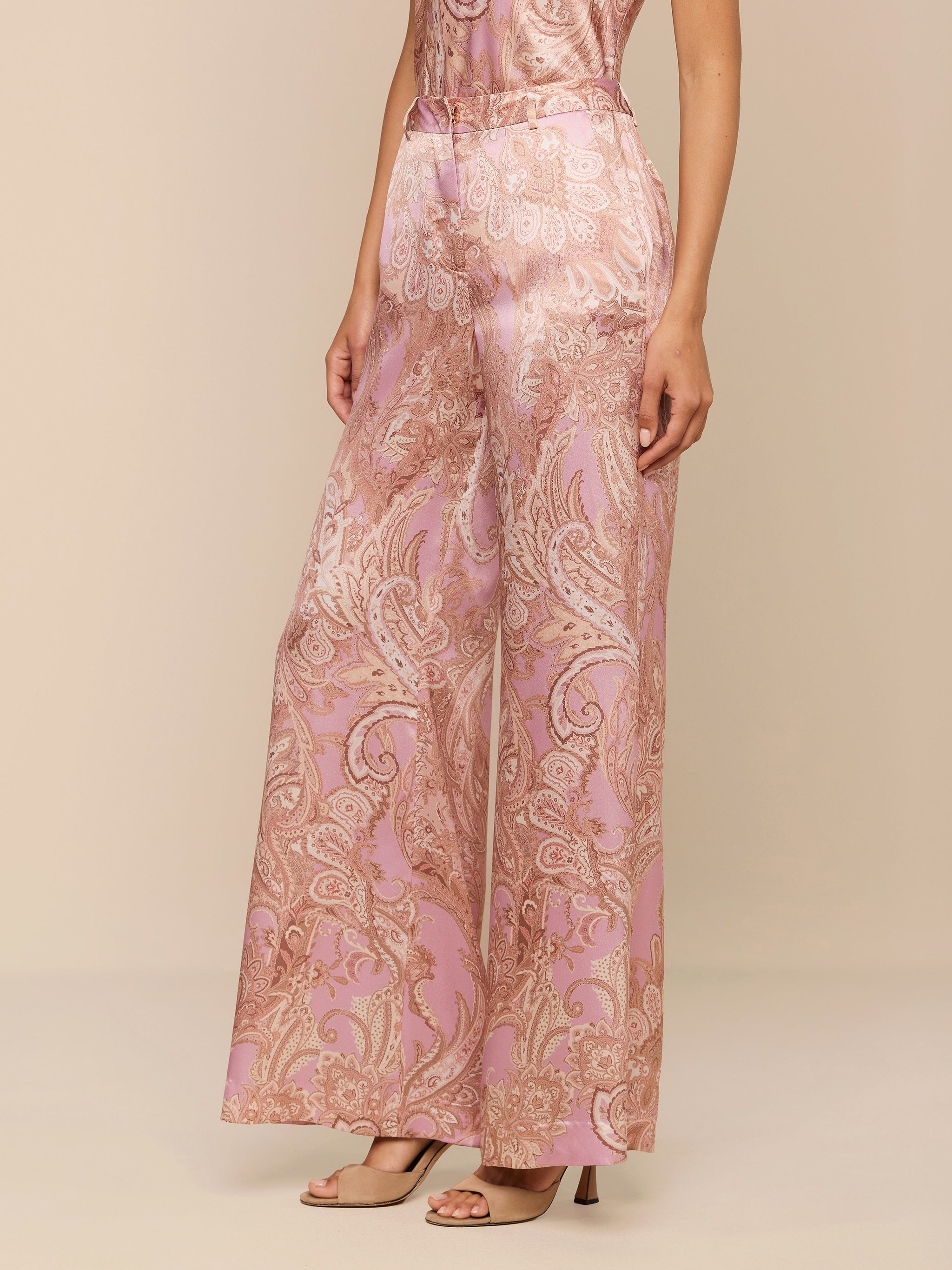 Portant le pantalon large Pilar en soie à imprimé cachemire rose et beige, associé à des talons nude ouverts, une personne se tient debout sur un fond beige, le haut de son corps étant partiellement hors champ.