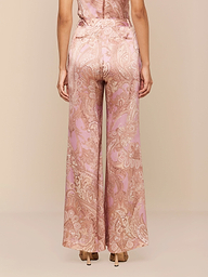 Une femme fait face à la caméra, vêtue dun pantalon large en soie Pilar à imprimé cachemire rose et or et de talons hauts beiges, sur un fond beige uni.