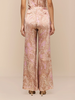 A woman faces away from the camera, wearing Pilar Silk Wide-Leg Pant in a pink and gold paisley print with beige high heels, set against a solid beige backdrop.