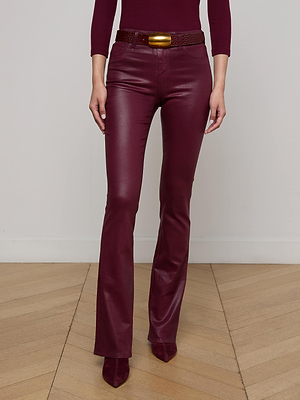 Wearing L'AGENCE Selma Coated Bootcut Jeans with a matching top, burgundy pointed-toe shoes, and a gold buckle belt, a person stands on a wooden floor against a plain light-colored wall.