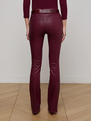 A person stands facing away from the camera, wearing L'AGENCE Selma Coated Bootcut Jean in maroon with a matching belt, on a light wood floor against a plain white wall.