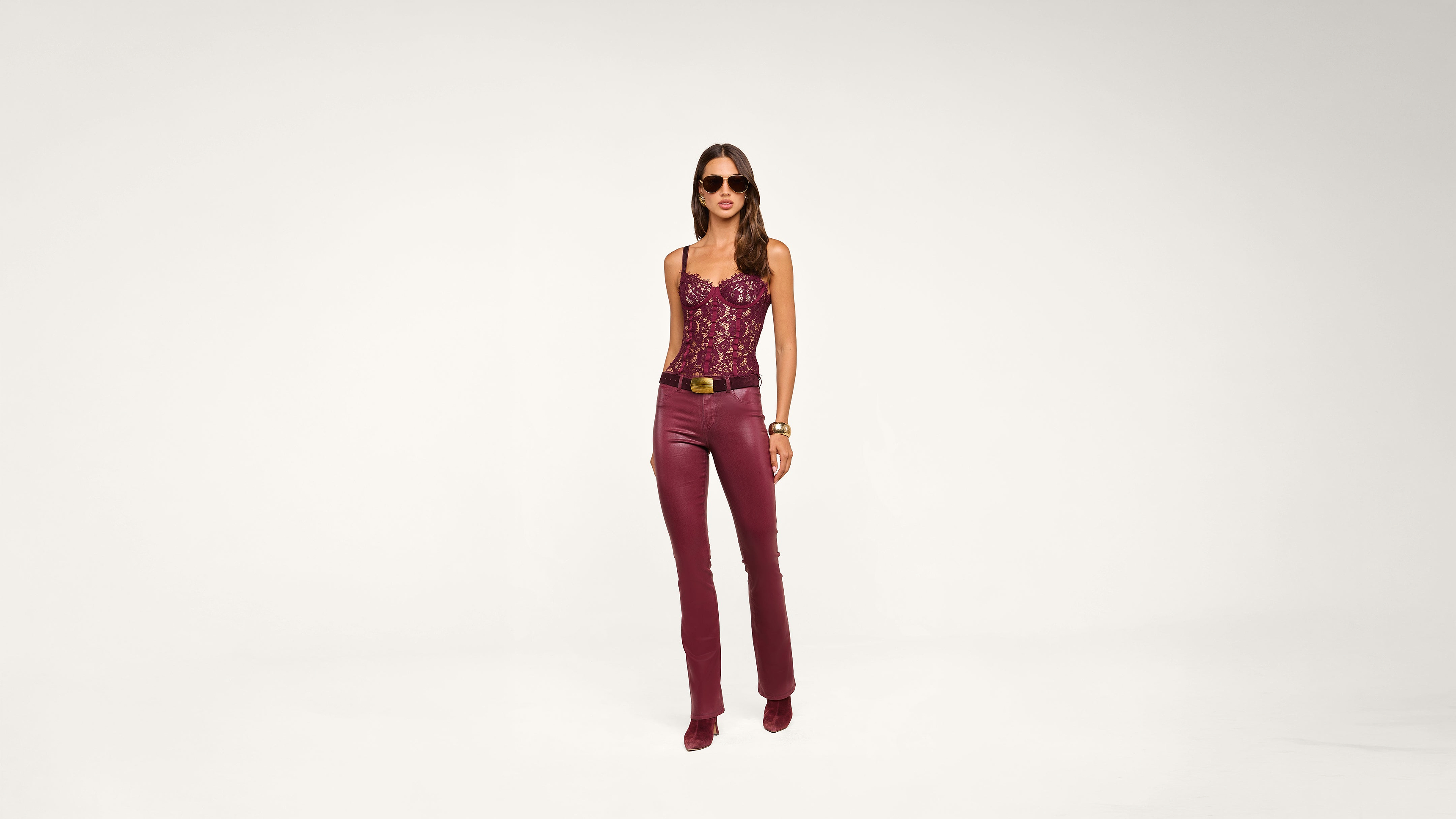 Woman wearing sunglasses, a maroon lace top, maroon leather pants, matching heels, a gold-buckled belt, and bracelets, standing against a plain white background.