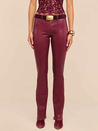 A woman wearing the Selma Coated Bootcut Jean in maroon, paired with a matching lace top, a black belt with a gold buckle, a gold bracelet, and burgundy shoes stands against a beige background.