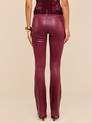 A person stands facing away, wearing the Selma Coated Bootcut Jean in shiny burgundy with a matching lace top, a gold bracelet on their wrist, and heels. The background is a plain beige color.