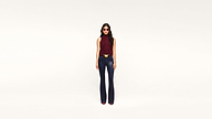 A woman wearing sunglasses, a sleeveless maroon top, dark pants, and red shoes stands confidently against a plain white background.